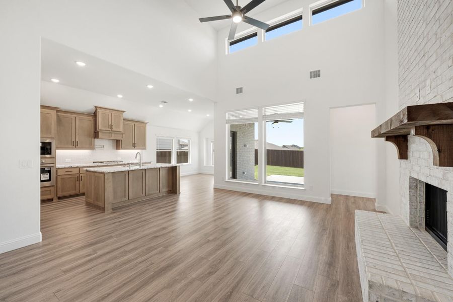 Open-concept living area with vaulted ceiling, wood-look floors, stone fireplace, and kitchen with island visible in background. Open-concept living area with vaulted ceiling, wood-look floors, stone fireplace, and kitchen with island visible in background.