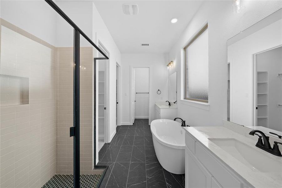 Spacious bathroom featuring a freestanding soaking tub, dual vanities with white countertops, a walk-in shower with light-toned tile, and dark-toned floor tiling