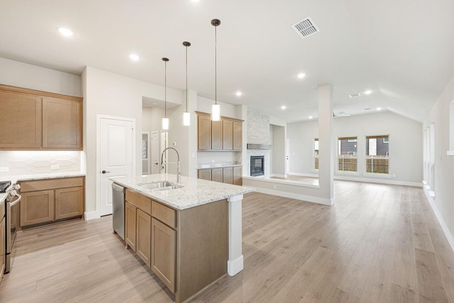 Open-concept kitchen with granite island, wood cabinets, and hardwood floors leading to living room with stone fireplace