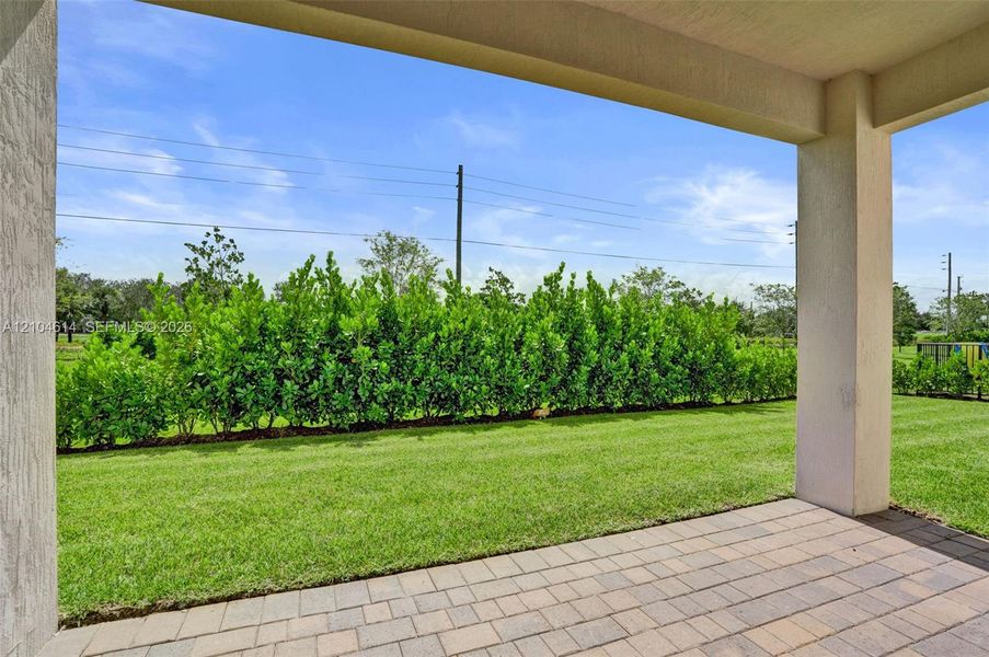 Expansive outdoor living area featuring a covered patio with paver flooring, lush lawn, and mature privacy hedging