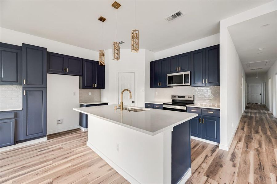 Kitchen featuring decorative backsplash, stainless steel appliances, a kitchen island with sink, pendant lighting, and light wood-type flooring