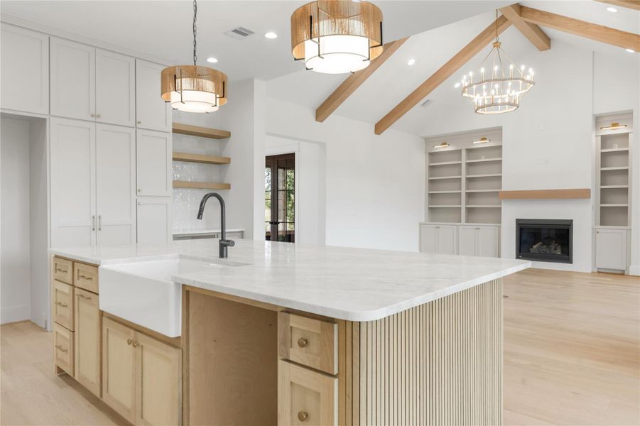 Dual tone kitchen featuring light wood-type flooring, a glass covered fireplace, a large island with sink, open floor plan, and beamed ceiling
