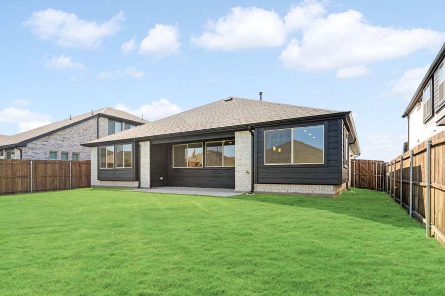 Back of single-story home with dark siding, stone accents, covered patio, and fenced green backyard