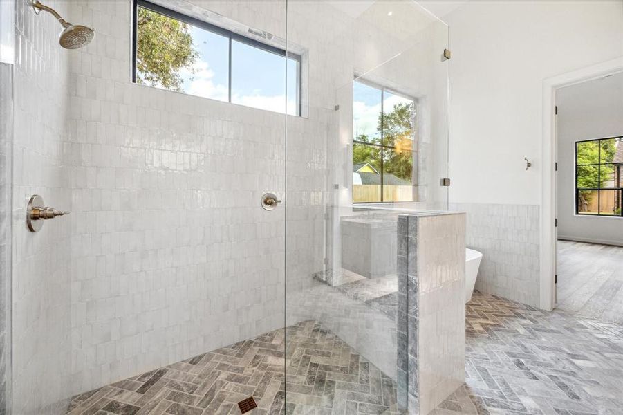 Luxurious shower area with frameless glass doors, tile walls up to the ceiling, 2 shower heads, bench in the shower and herringbone-patterned flooring, highlighted by large windows providing natural light.