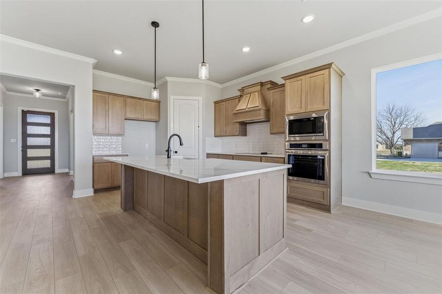 Kitchen featuring appliances with stainless steel finishes, a kitchen island with sink, decorative light fixtures, decorative backsplash, and premium range hood
