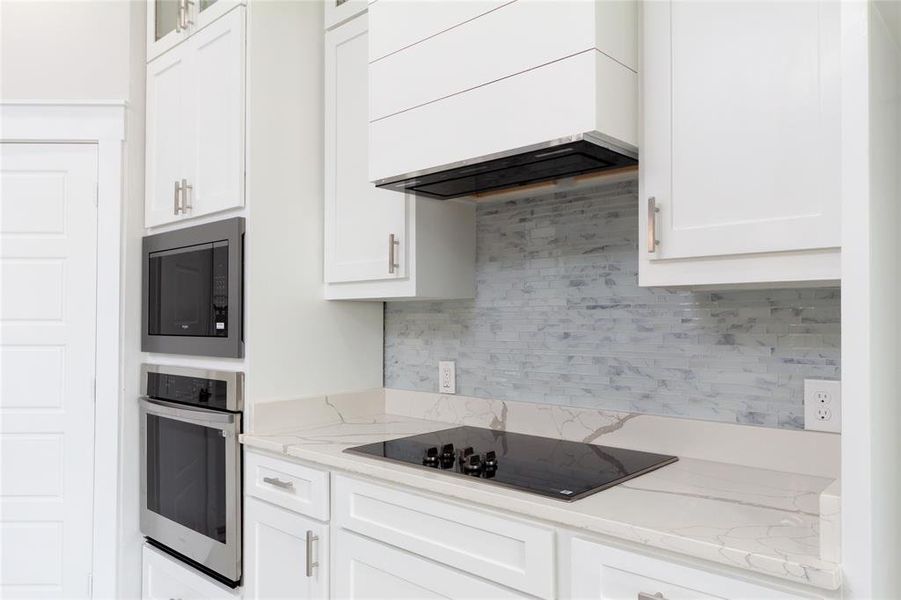 Kitchen with tasteful backsplash, light stone counters, custom range hood, stainless steel appliances, and white cabinets