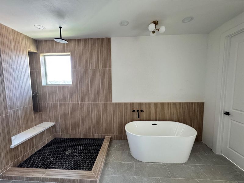 This bathroom features a modern design with a spacious walk-in shower, sleek black hexagonal tiles, and a rainfall showerhead. It also includes a stylish freestanding bathtub and contemporary light fixtures, set against wood-accented walls and a neutral color palette.