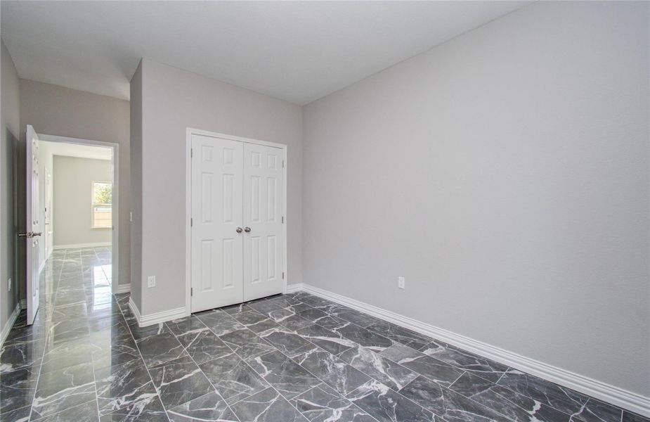 This photo shows a room with sleek, dark marble tile flooring and neutral gray walls. There's a double-door closet and a hallway leading to a sunlit space with a window. The overall look is modern and clean.