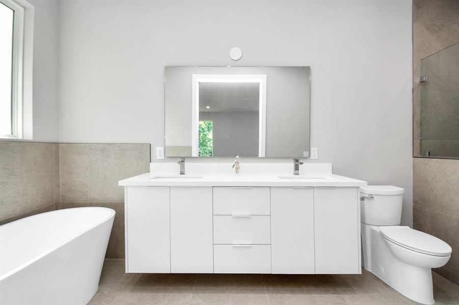 This is a modern bathroom featuring a standalone bathtub, a floating vanity with a large mirror, and a toilet. The space has neutral colors and large tiles, with ample natural light from the frosted window.