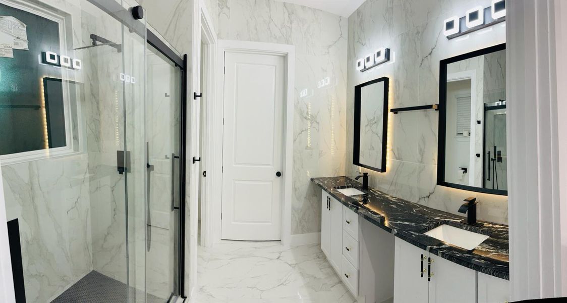 Full bathroom with double vanity, a marble finish shower, stone wall, and light marble finish floors Full bathroom with double vanity, a marble finish shower, stone wall, and light marble finish floors