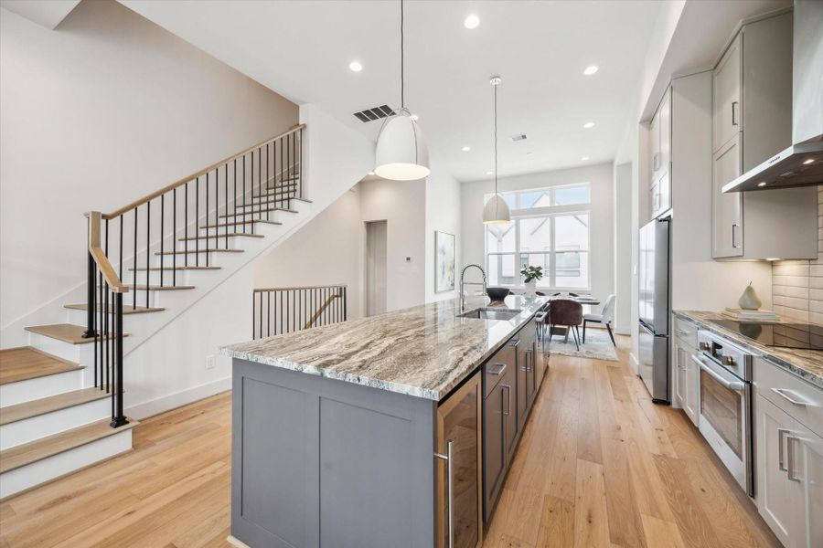 This modern kitchen features a large, elegant island with marble countertops, stainless steel appliances, and light wood flooring. It opens to a bright dining area with large windows, providing ample natural light. The space is complemented by a stylish staircase and contemporary pendant lighting.
