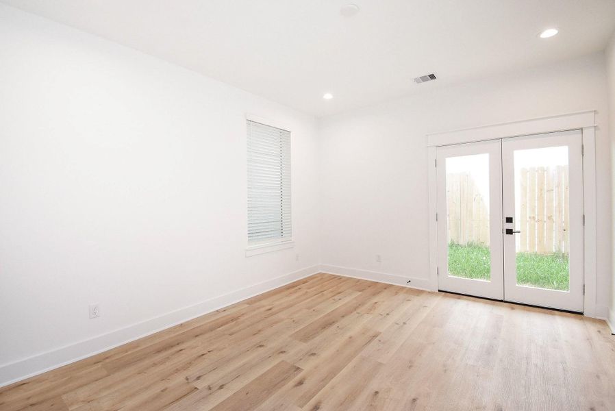 Bright room with wood flooring, white walls, and recessed lighting. It features a window with blinds and French doors leading to a fenced yard.