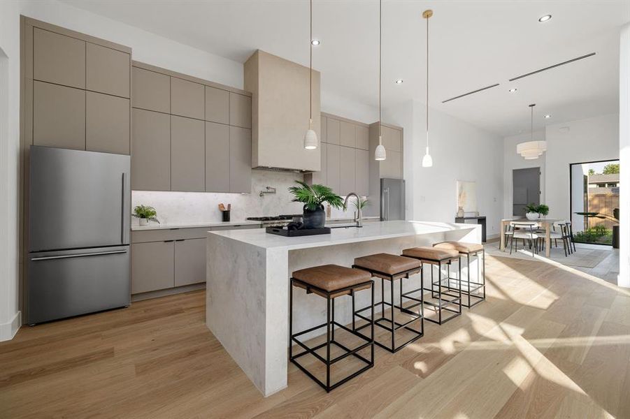 Modern kitchen featuring light wood-finish flooring, full-height cabinetry with integrated refrigerator, a large center island with waterfall countertop, and pendant lighting