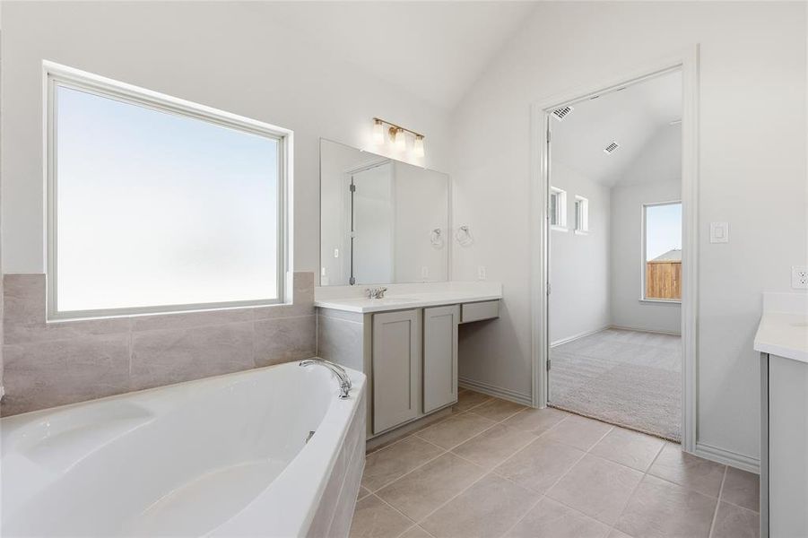 Soaking tub with tiled surround and an expansive window, complemented by a dual vanity with light gray cabinetry and a separate vanity area