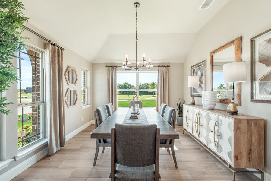 Dining room with long table, chandelier, vaulted ceiling, and large windows overlooking a backyard