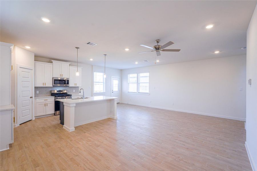 This photo showcases a spacious open-concept living area with light wood flooring and neutral walls. The modern kitchen features white cabinetry, stainless steel appliances, and a central island with pendant lighting. The room is brightened by large windows and recessed lighting, and a ceiling fan adds comfort.
