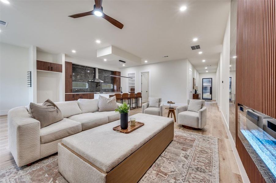 Open-concept living area with light wood-finish flooring, recessed lighting, a modern ceiling fan, a built-in electric fireplace with slatted wood surround, and a kitchen featuring dark stone backsplash and countertops