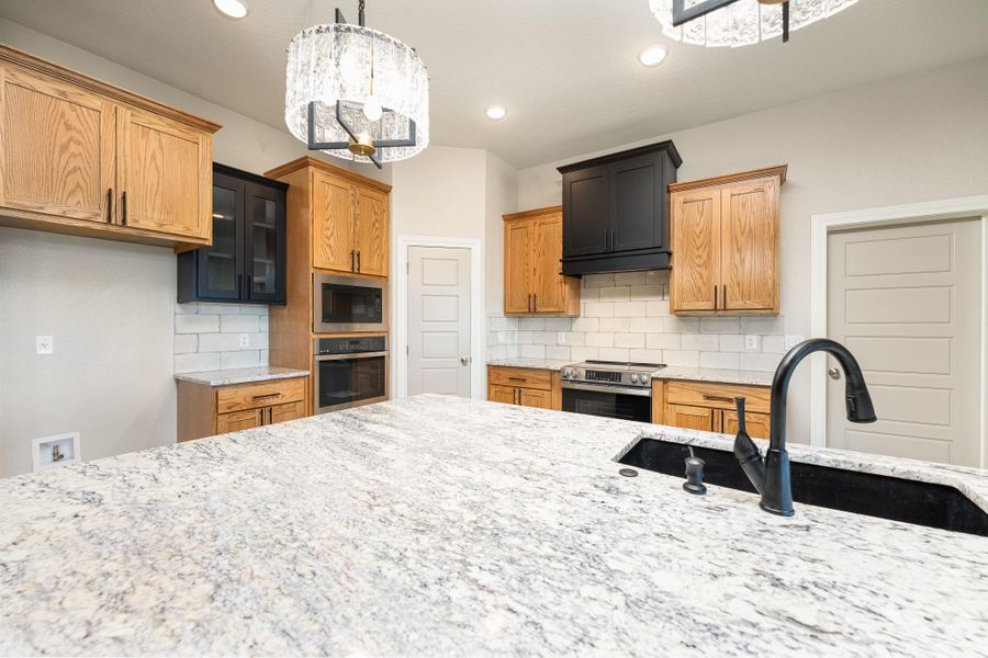 Kitchen featuring decorative backsplash, brown cabinetry, light stone counters, a chandelier, and stainless steel appliances Kitchen featuring decorative backsplash, brown cabinetry, light stone counters, a chandelier, and stainless steel appliances