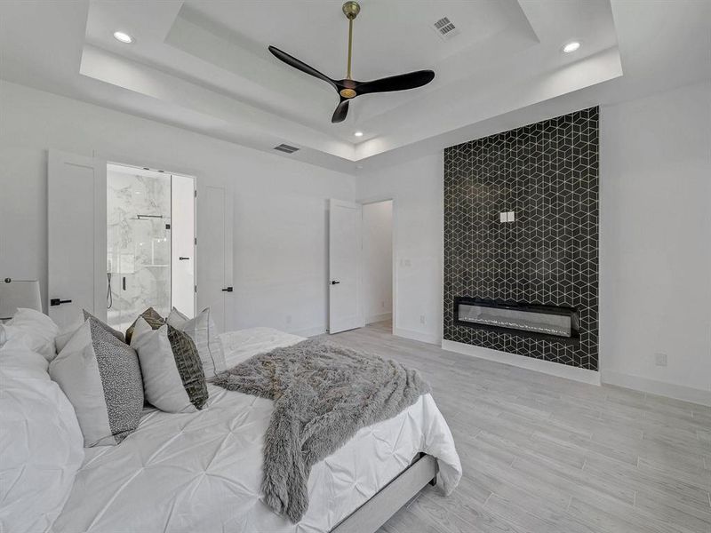 Bedroom featuring a tray ceiling, a tiled fireplace, recessed lighting, light wood-type flooring, and connected bathroom