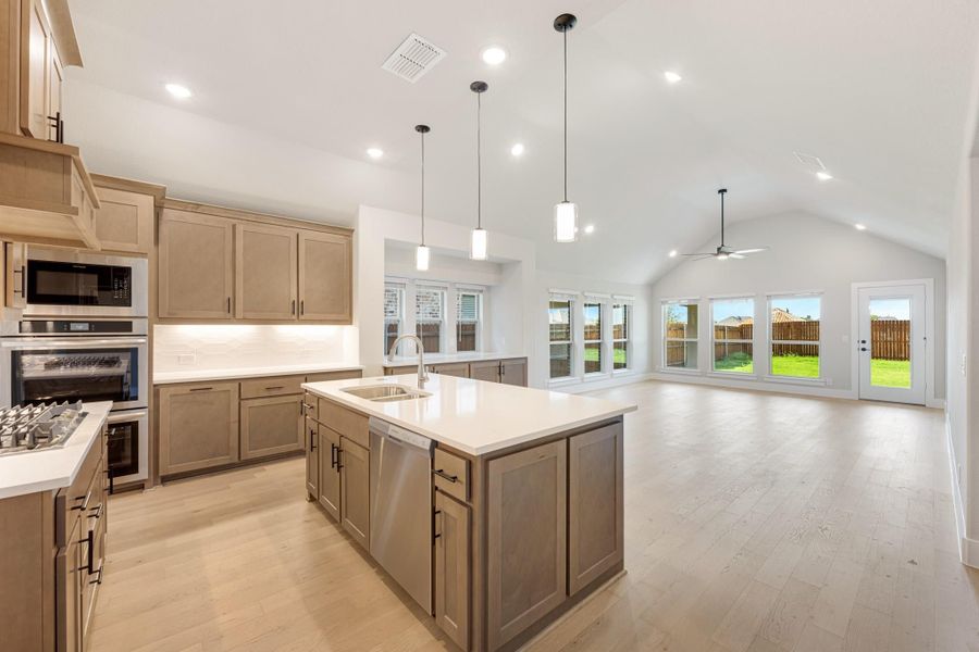 Open kitchen with large island, tan cabinets, stainless appliances, and vaulted ceiling open to living area with backyard views Open kitchen with large island, tan cabinets, stainless appliances, and vaulted ceiling open to living area with backyard views