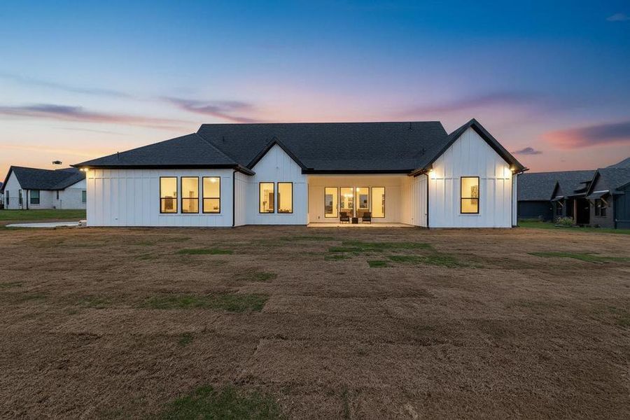 Back of house at dusk featuring a patio, board and batten siding, roof with shingles, and a yard Back of house at dusk featuring a patio, board and batten siding, roof with shingles, and a yard