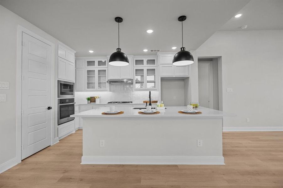 Spacious kitchen featuring an expansive central island with seating, pendant lighting, white cabinetry, integrated stainless steel appliances, and wood-finish flooring Spacious kitchen featuring an expansive central island with seating, pendant lighting, white cabinetry, integrated stainless steel appliances, and wood-finish flooring