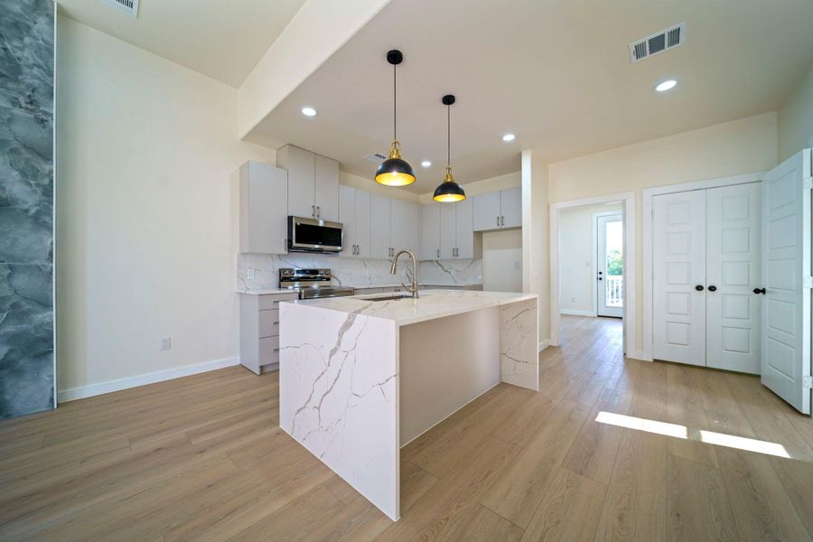 This modern kitchen features sleek white cabinetry, a spacious quartz island with waterfall edges, and stylish pendant lighting. The open layout is complemented by light LVP flooring and stainless steel appliances, creating a bright, inviting space. This modern kitchen features sleek white cabinetry, a spacious quartz island with waterfall edges, and stylish pendant lighting. The open layout is complemented by light LVP flooring and stainless steel appliances, creating a bright, inviting space.