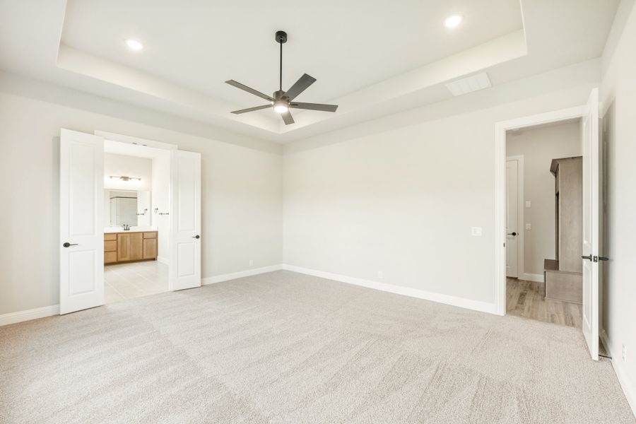 Spacious master bedroom with tray ceiling, ceiling fan, carpet flooring, and open doors to ensuite bathroom and hallway. Spacious master bedroom with tray ceiling, ceiling fan, carpet flooring, and open doors to ensuite bathroom and hallway.