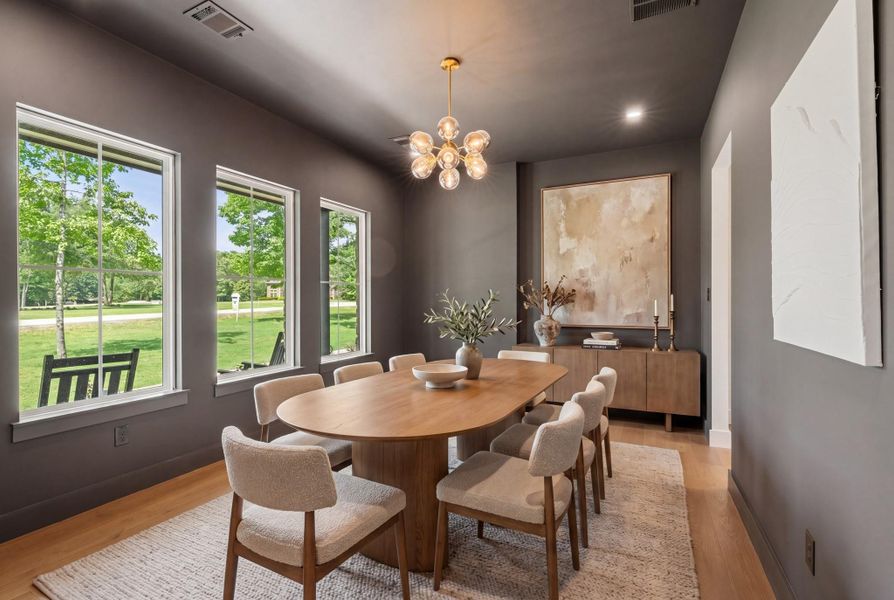 Formal dining room with dramatic charcoal walls, warm 7" wood plank flooring and abundant natural light from three oversized windows. A modern brass chandelier creates a bold focal point overhead. Open to the adjoining living and kitchen area, this space is well-suited for hosting dinners and family gatherings.