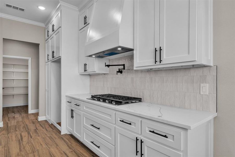 Kitchen featuring wall chimney range hood, white cabinetry, wood finished floors, decorative backsplash, and crown molding Kitchen featuring wall chimney range hood, white cabinetry, wood finished floors, decorative backsplash, and crown molding