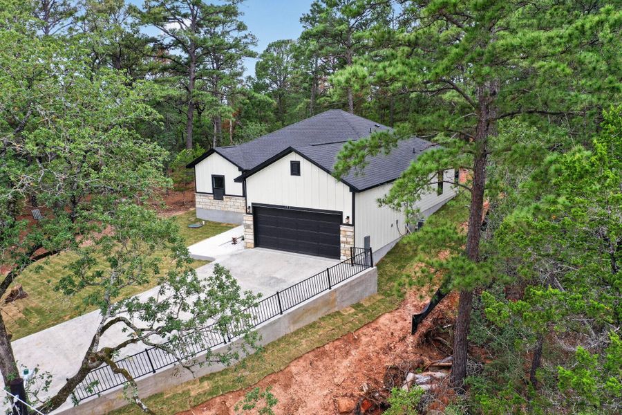 Modern farmhouse-style exterior featuring white vertical siding, stone accents, a dark shingle roof, an attached garage with a black door, and a concrete driveway with a black metal railing Modern farmhouse-style exterior featuring white vertical siding, stone accents, a dark shingle roof, an attached garage with a black door, and a concrete driveway with a black metal railing