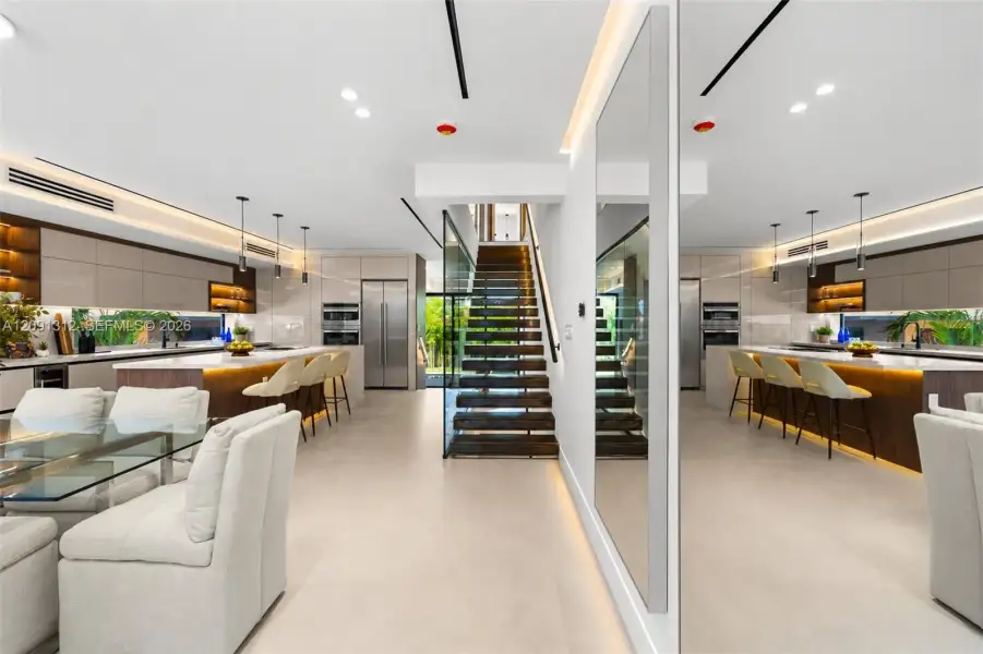 Modern open-concept area featuring sleek flat-panel cabinetry, a central island with integrated lighting, polished stone countertops, and a staircase with wood-tread risers and glass balustrades Modern open-concept area featuring sleek flat-panel cabinetry, a central island with integrated lighting, polished stone countertops, and a staircase with wood-tread risers and glass balustrades