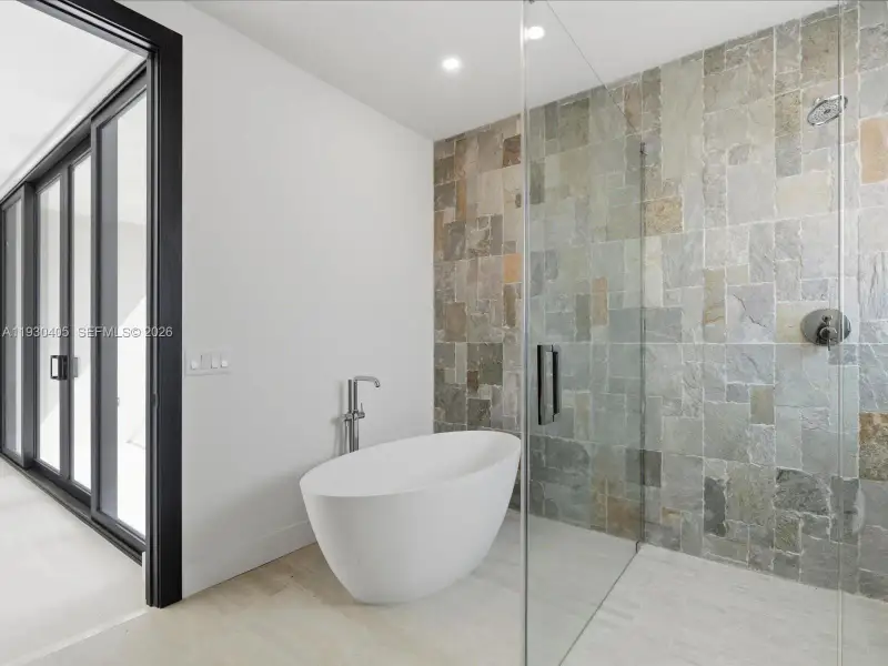 Freestanding white soaking tub with a floor-mounted faucet, glass-enclosed walk-in shower with natural stone tile wall, and sliding glass doors with black framing Freestanding white soaking tub with a floor-mounted faucet, glass-enclosed walk-in shower with natural stone tile wall, and sliding glass doors with black framing