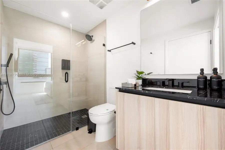 Bathroom featuring a floating vanity with a dark-toned stone countertop, walk-in shower with a glass enclosure and handheld shower head, and large-format wall tiling Bathroom featuring a floating vanity with a dark-toned stone countertop, walk-in shower with a glass enclosure and handheld shower head, and large-format wall tiling
