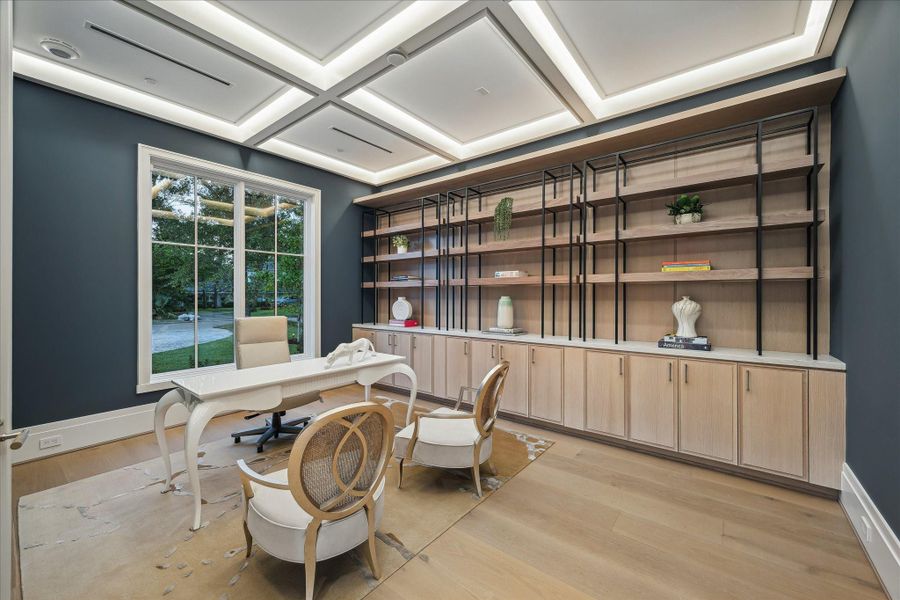 Stylish home office library featuring a striking coffered ceiling with recessed lighting and crisp trim detailing. A full wall of custom built-ins includes light oak cabinetry topped with stone surfaces and open shelving framed in sleek black metal. Large windows bring in abundant natural light, while wide-plank oak flooring and deep-toned accent walls create a polished workspace.