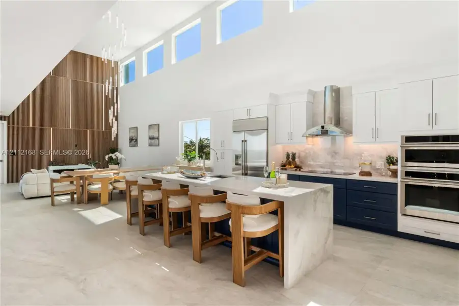 Open-concept kitchen featuring double-height ceilings, a large stone-topped waterfall island, custom two-toned cabinetry, built-in stainless steel appliances, and a floor-to-ceiling wood panel accent wall Open-concept kitchen featuring double-height ceilings, a large stone-topped waterfall island, custom two-toned cabinetry, built-in stainless steel appliances, and a floor-to-ceiling wood panel accent wall