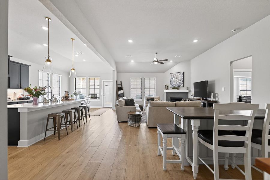 This open-concept living space features a modern kitchen with a large island and bar seating, flowing into a spacious living room with a cozy fireplace. The room is brightened by ample natural light from large windows, complemented by stylish vinyl wood floors and neutral tones.