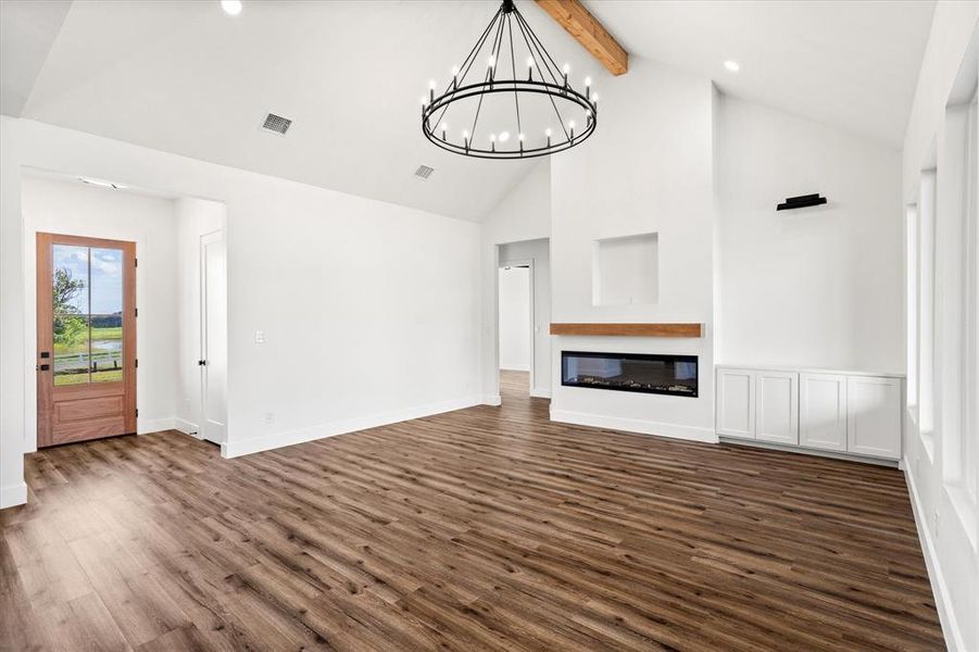 Unfurnished living room featuring dark wood-type flooring, high vaulted ceiling, a glass covered fireplace, beamed ceiling, and a chandelier Unfurnished living room featuring dark wood-type flooring, high vaulted ceiling, a glass covered fireplace, beamed ceiling, and a chandelier