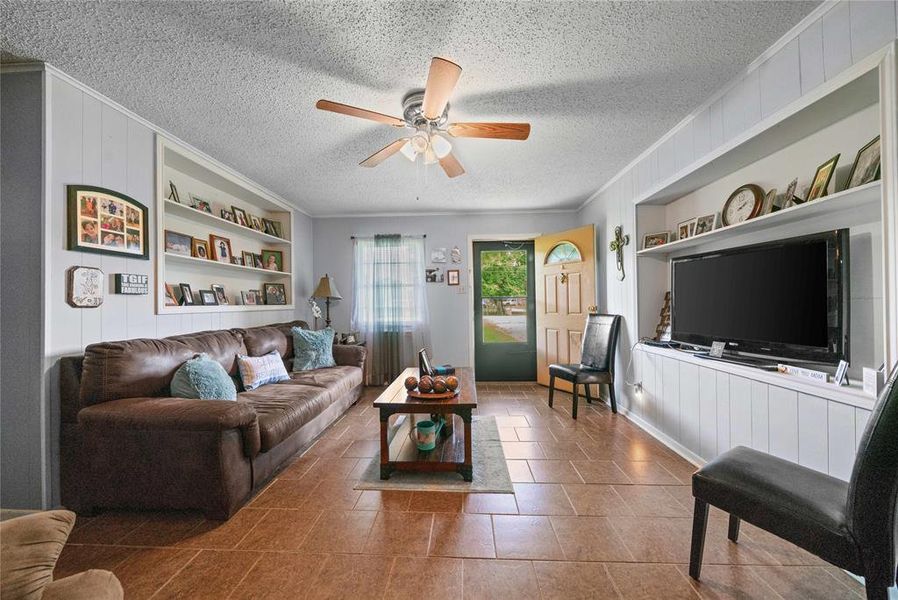 Living area with built in features, ornamental molding, a textured ceiling, a ceiling fan, and wood walls