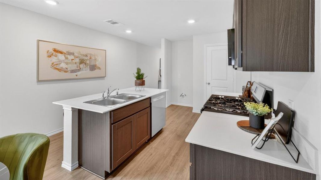 Open-concept kitchen featuring an island with a double basin stainless steel sink, wood-finish flooring, recessed lighting, white countertops, and dark wood-finish cabinetry Open-concept kitchen featuring an island with a double basin stainless steel sink, wood-finish flooring, recessed lighting, white countertops, and dark wood-finish cabinetry