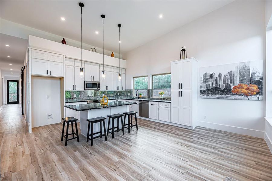 The modern kitchen features white cabinetry, a large island with seating, stainless steel appliances, and stylish pendant lighting, and set against a backdrop of light vinyl floors.