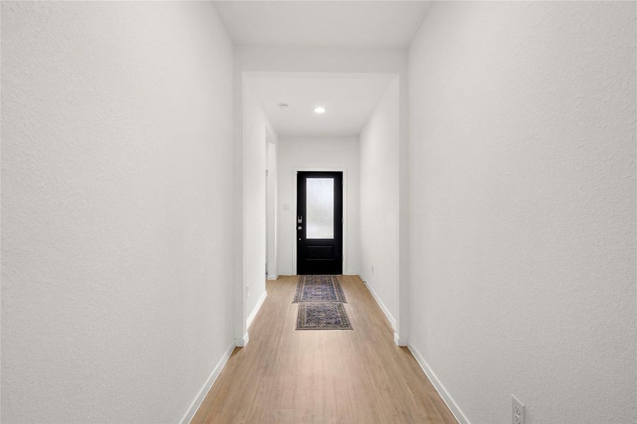 This photo shows a bright, minimalistic hallway with light wood flooring and white walls. It features a black front door with frosted glass, creating a modern and welcoming entryway. A narrow rug adds a subtle touch of warmth.