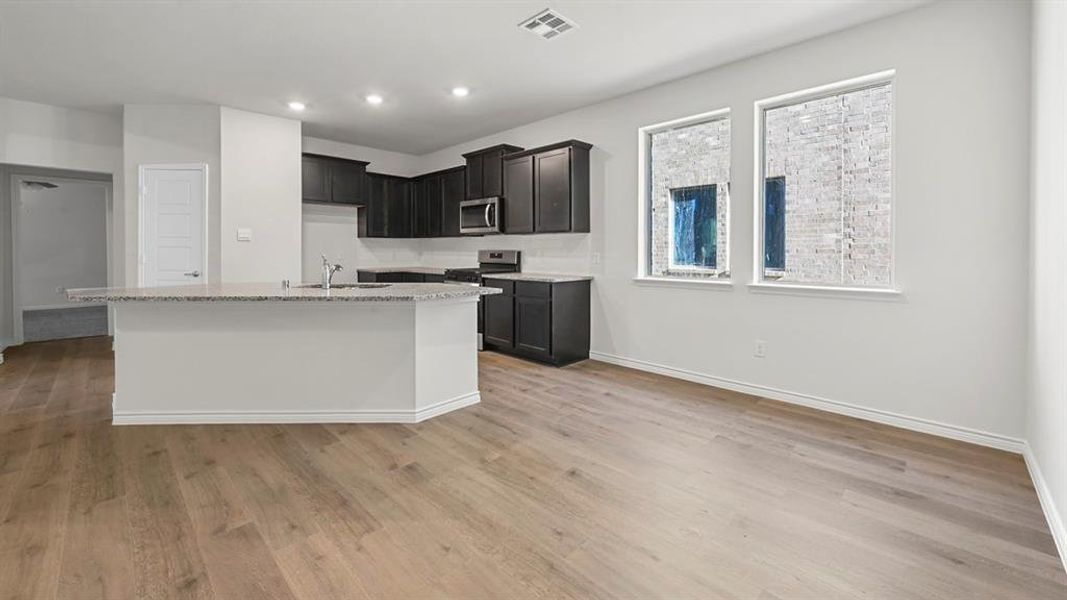Kitchen featuring light stone counters, dark cabinets, light wood-type flooring, stainless steel appliances, and a kitchen island with sink