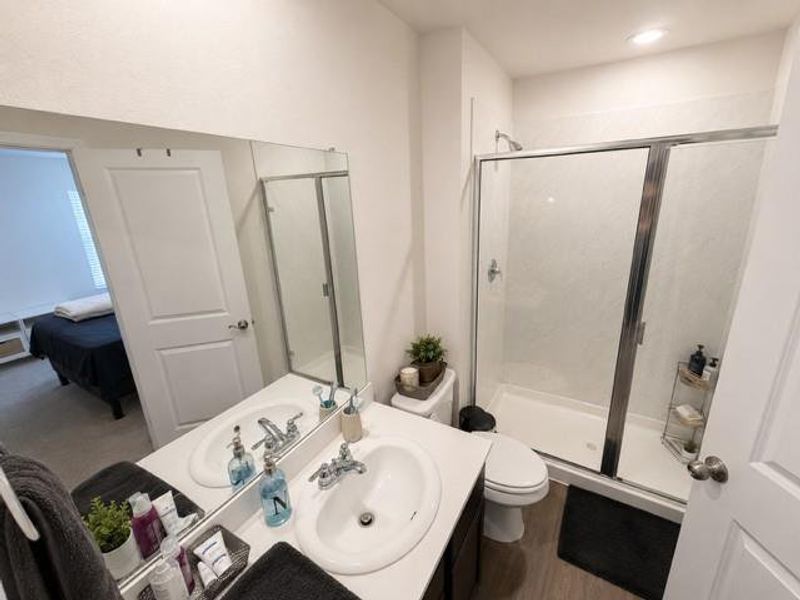 Bathroom featuring a white vanity with dual sinks, a large mirror, a glass-enclosed shower with tiled walls, and wood-finish flooring Bathroom featuring a white vanity with dual sinks, a large mirror, a glass-enclosed shower with tiled walls, and wood-finish flooring