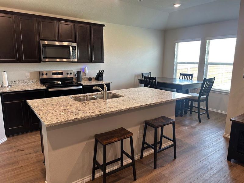 Kitchen with dark brown cabinets, appliances with stainless steel finishes, dark wood-type flooring, light stone countertops, and an island with sink
