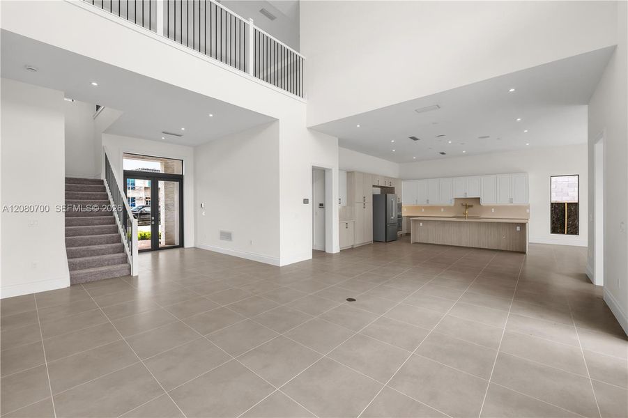 Open-concept living space featuring high ceilings, large format tile flooring, a staircase with carpeted treads and metal balusters, and a kitchen with light wood-finish cabinetry and a built-in refrigerator