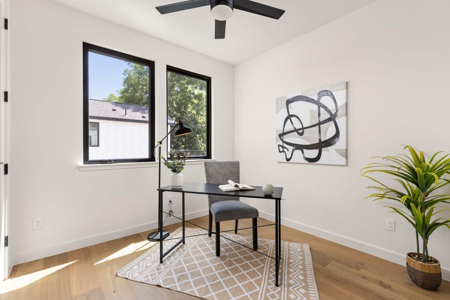 A well-lit study with ample natural light, featuring elegant hardwood flooring and a contemporary ceiling fan.