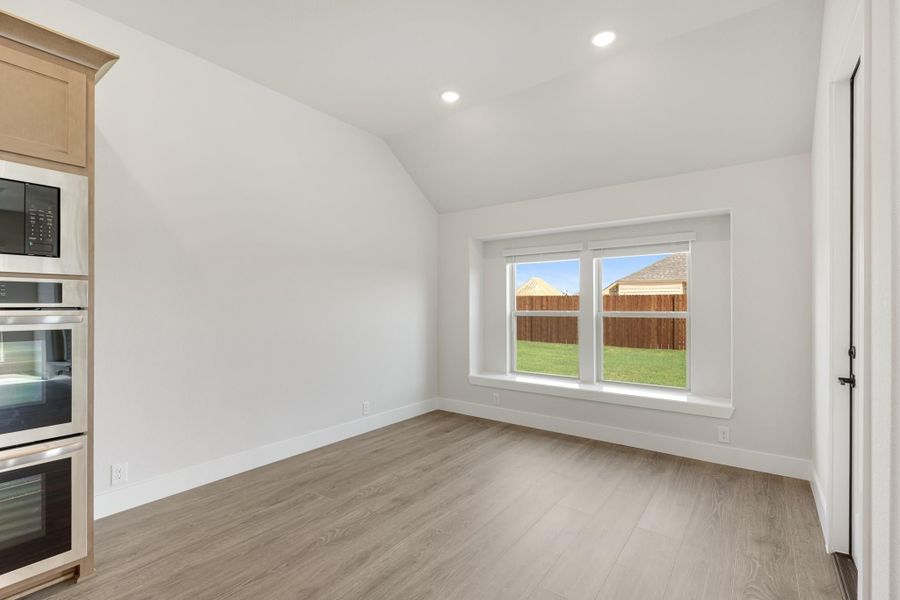 Empty dining area with vaulted ceiling, wood-look floors, and backyard-facing window beside built-in ovens