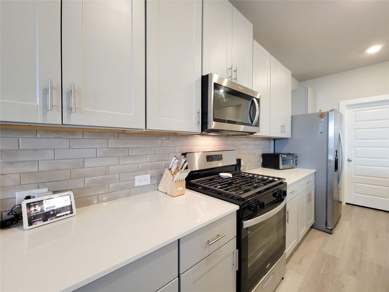 This modern kitchen features sleek white cabinetry, stainless steel appliances, a gas stove, and a stylish subway tile backsplash. The space is well-lit and includes ample countertop area for cooking and entertaining.