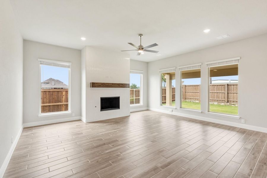 Open living room with white tile fireplace, wood-look floors, ceiling fan, and large windows overlooking backyard